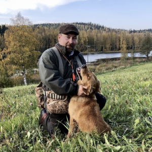 Mann mit Hund beim Spaziergang im Grünen, Natur und Wasser im Hintergrund.